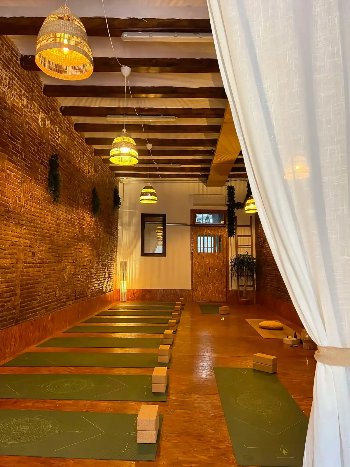 Sala grande con esterillas verdes en fila, bloques de corcho, pared de ladrillo y lámparas de mimbre