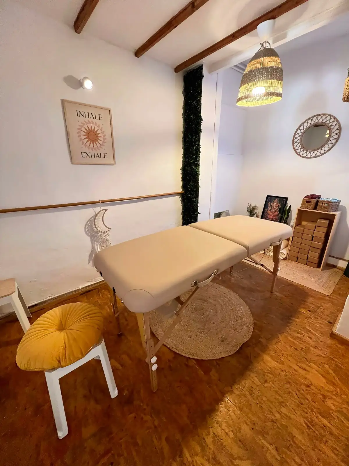 Sala pequeña con camilla de masaje, taburete, alfombra de yute y lámina «Inhale · Exhale» en la pared