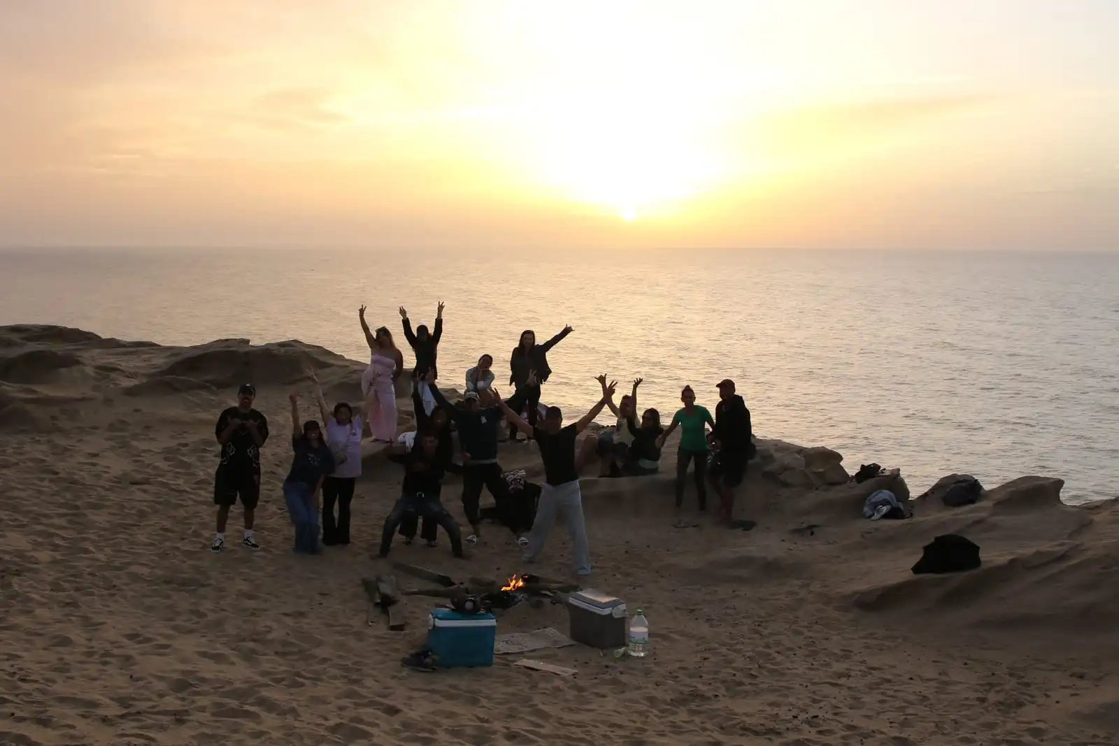 Atardecer sobre el océano desde las dunas, con el grupo celebrando en la arena